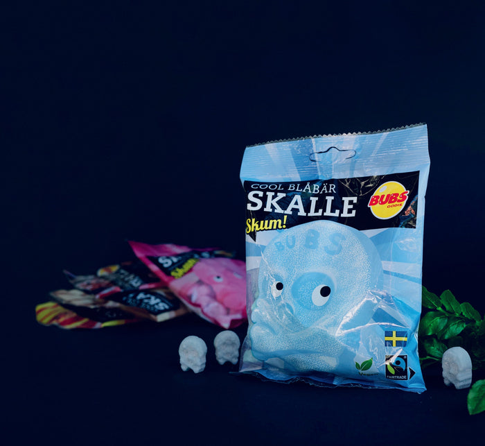 Three bags of BUBS Skull candy against a black background, featuring a focus on the small blue candy skulls scattered around the bags. The bags showcase the Cool Blueberry flavored candy, highlighting the vibrant blue hues and playful design.